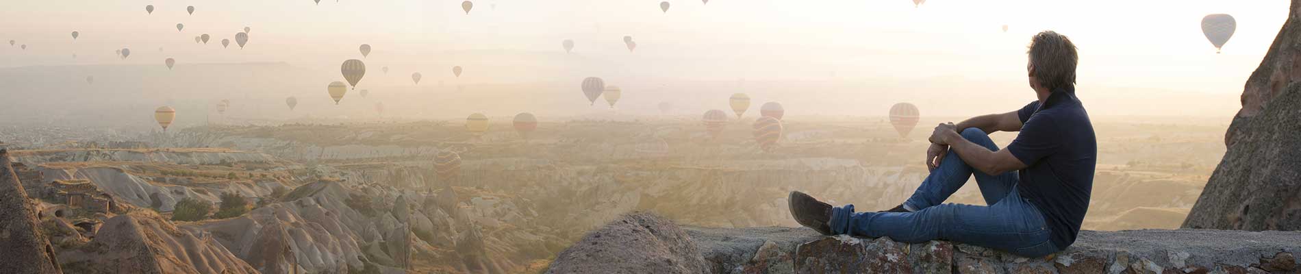 A man sitting on a rock plateau watching hot air balloons take to flight