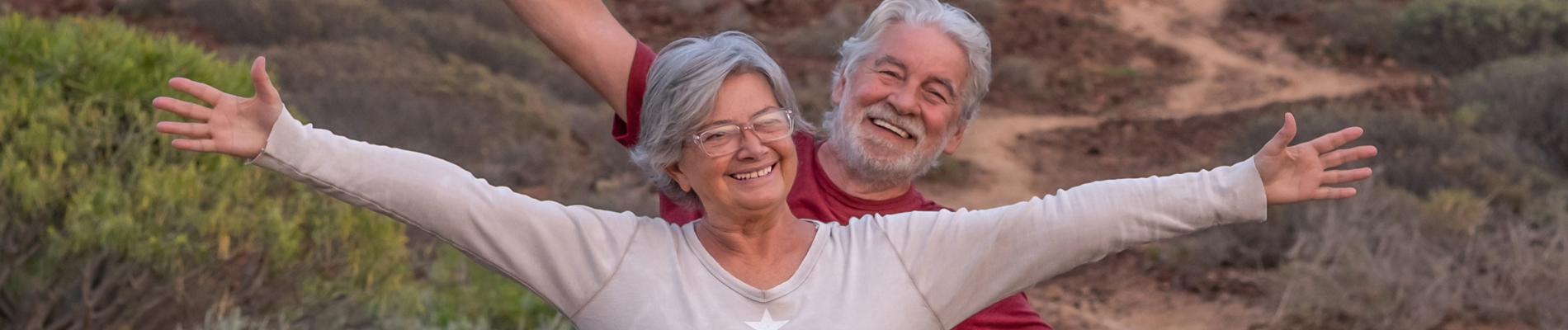 older couple grinning with out stretched arms on a hiking trail