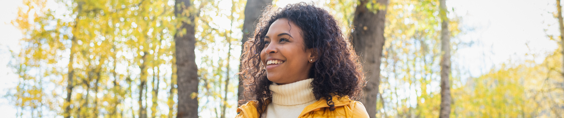 Woman with a white turtle neck and yellow coat smiling in the woods