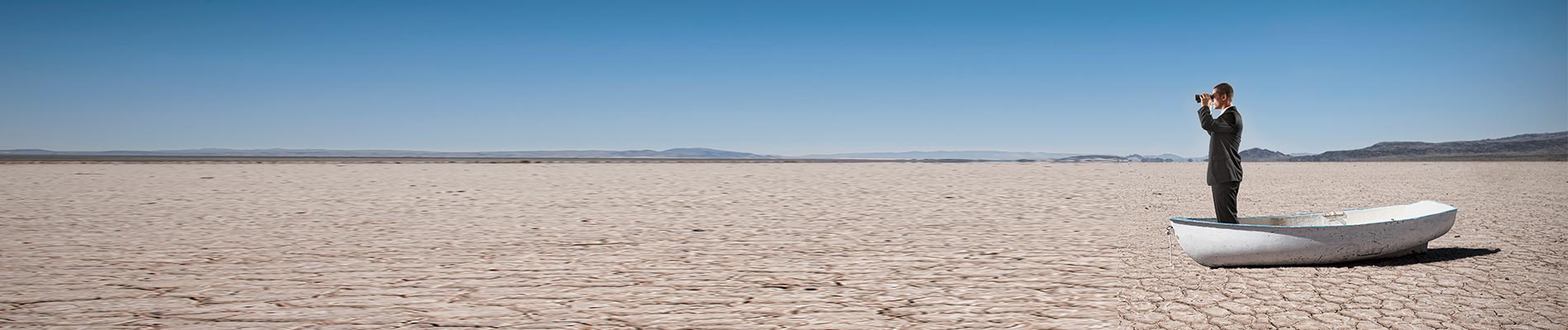 A man in a suit looking through field glasses standing in a boat on a dry lake bed