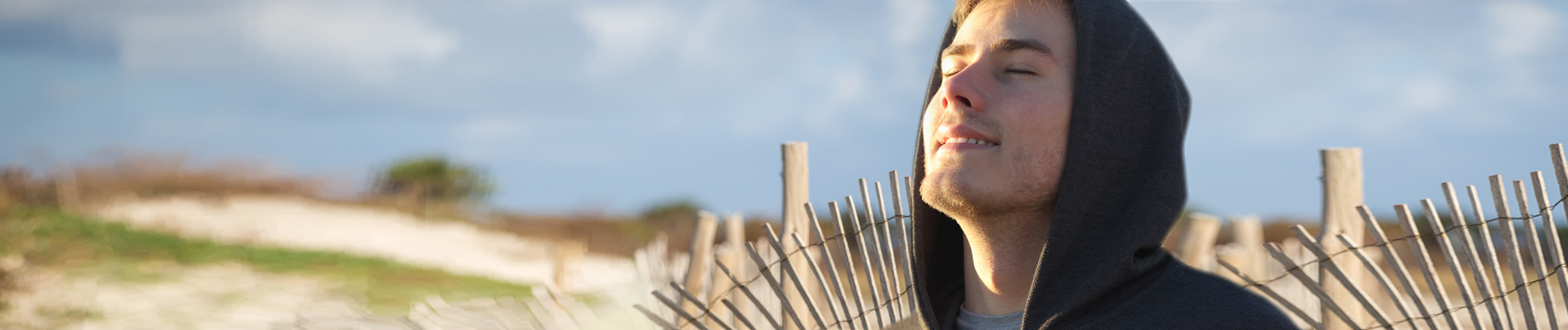 A man in a hoody, head tilted up into the sunlight next to a fence along sand dunes